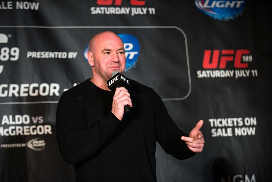 Dana White w czarnym swetrze stoi na tle baneru konferencyjnego UFC