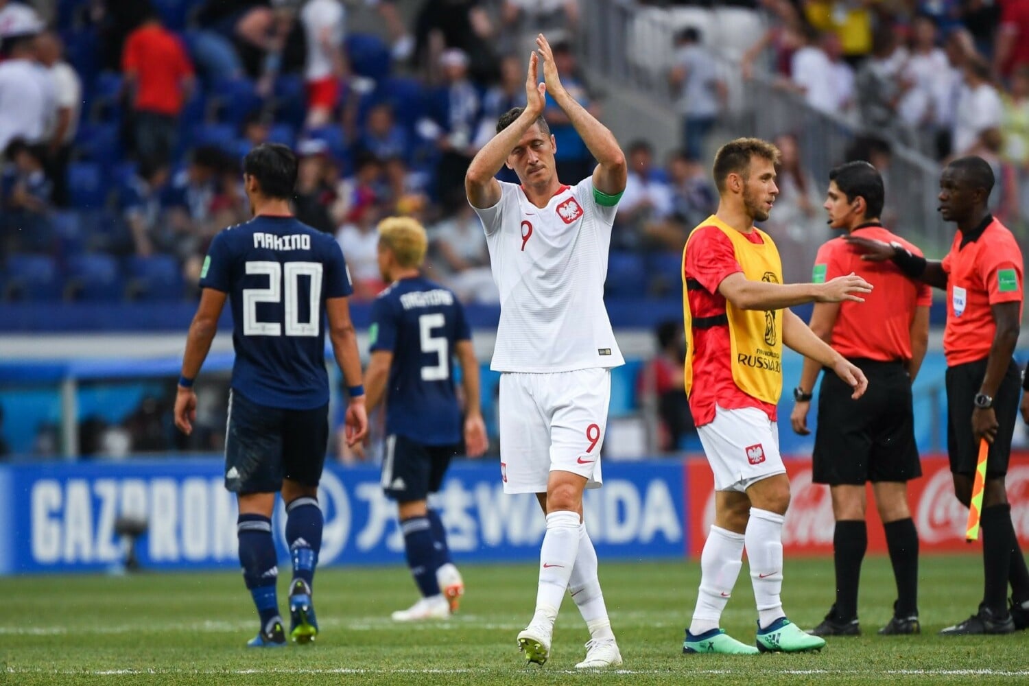 Zdjęcie Roberta Lewandowskiego z meczu z Japonią z 2018 roku. Kapitan reprezentacji Polski schodzi z murawy i żegna się z kibicami. W tle zawodnicy reprezentacji Japonii w granatowych trykotach.