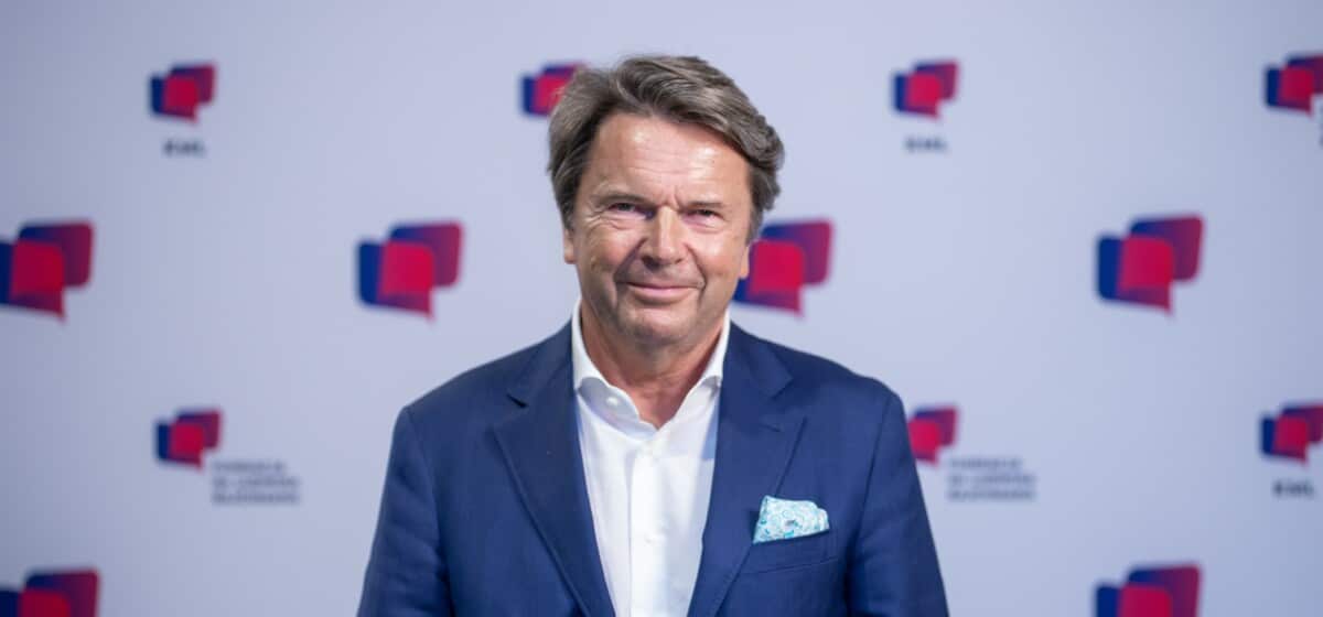 Zbigniew Jakubas pozuje do zdjęcia na tle ścianki. Ma na sobie niebiesko-granatową marynarkę i białą koszulę.