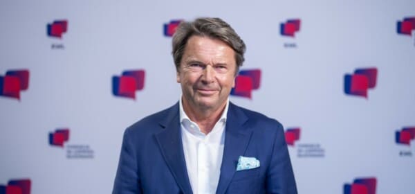 Zbigniew Jakubas pozuje do zdjęcia na tle ścianki. Ma na sobie niebiesko-granatową marynarkę i białą koszulę.