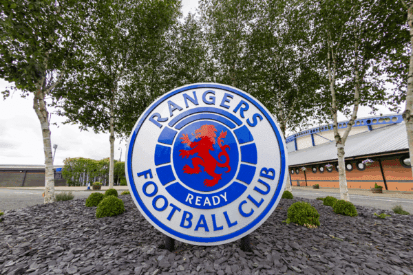 Duże, okrągłe logo Rangers Football Club z czerwonym lwem na niebieskim tle, ustawione przed klubowym obiektem i otoczone drzewami.