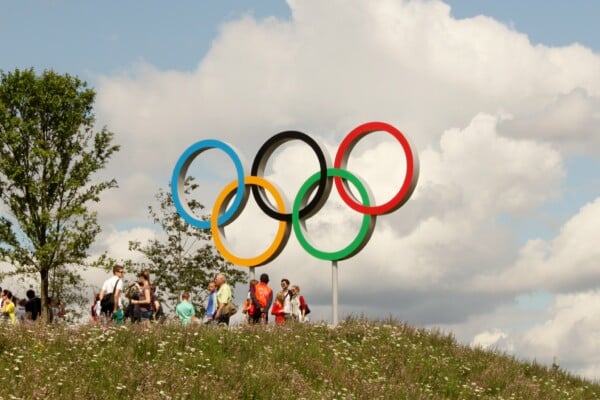 Grupa ludzi spaceruje po łące pod dużymi pierścieniami olimpijskimi na tle nieba i chmur