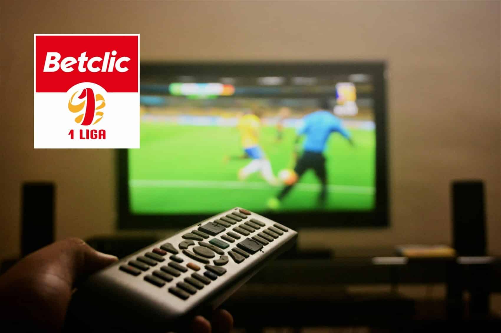 Osoba trzymająca pilot przed telewizorem, na którym wyświetlany jest mecz piłki nożnej.