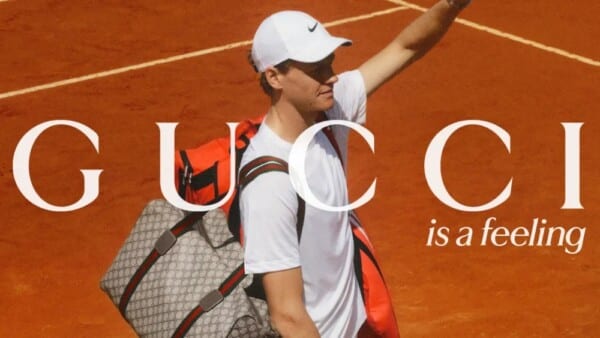 Jannik Sinner na korcie ziemnym niesie dużą torbę sportową Gucci oraz plecak, ubrany w białą koszulkę i czapkę z daszkiem, z uniesioną ręką w geście powitania lub triumfu. Na zdjęciu widnieje napis „GUCCI is a feeling”.