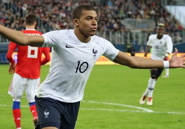 Kylian Mbappé w koszulce reprezentacji Francji z numerem 10 celebruje zdobycie bramki podczas meczu piłkarskiego na stadionie, w tle zawodnicy drużyny przeciwnej.