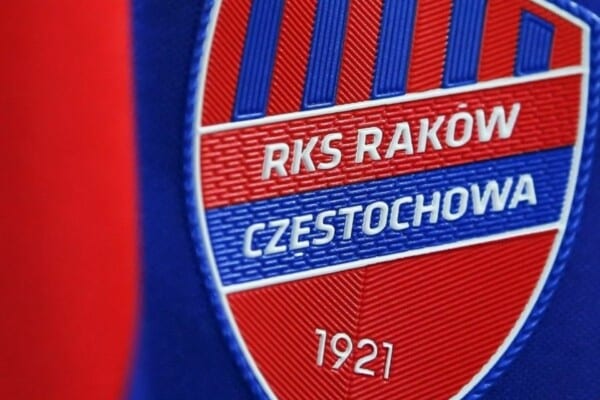 Herb Rakowa Częstochowa w kształcie tarczy, w czerwono-niebieskich barwach, z białym napisem „RKS Raków Częstochowa 1921”.