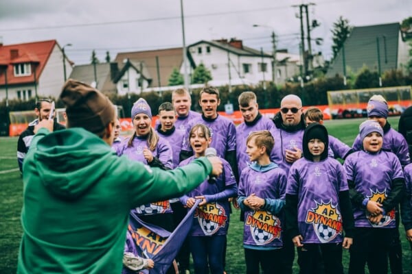 Zdjęcie przedstawia trenera i grupę młodych zawodników drużyny AP Dynamit w trakcie wydarzenia piłkarskiego Futbol Plus. Uczestnicy ubrani są w charakterystyczne fioletowe stroje z logo drużyny. Spotkanie odbywa się na zielonym boisku, a w tle widać zabudowania i elementy infrastruktury sportowej.