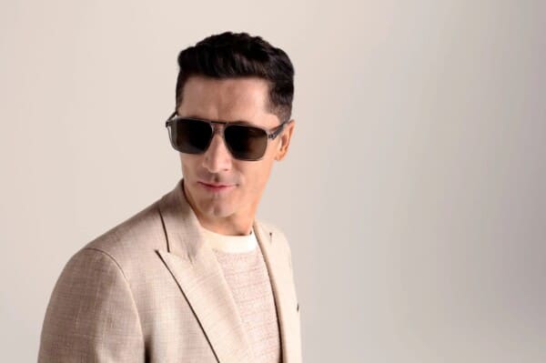 Robert Lewandowski prezentuje się w eleganckim, jasnym garniturze oraz modnych okularach z kolekcji BOSS Eyewear na sezon wiosna/lato 2025. Zdjęcie wykonano na tle jednolitej, jasnej ściany, co podkreśla nowoczesny i stylowy charakter wizerunku piłkarza.
