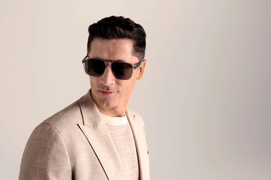 Robert Lewandowski prezentuje się w eleganckim, jasnym garniturze oraz modnych okularach z kolekcji BOSS Eyewear na sezon wiosna/lato 2025. Zdjęcie wykonano na tle jednolitej, jasnej ściany, co podkreśla nowoczesny i stylowy charakter wizerunku piłkarza.
