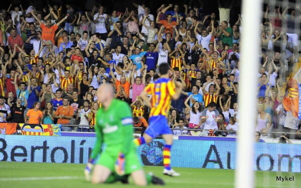 Tłum kibiców Valencii w żółto-czerwonych barwach świętuje bramkę na stadionie Mestalla podczas meczu piłkarskiego, piłkarz w pasiastym stroju biegnie w stronę trybun, a bramkarz na pierwszym planie klęczy na murawie.