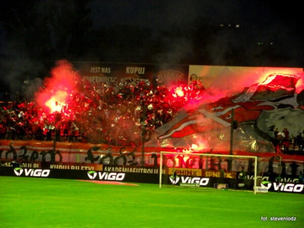 Stadion Widzewa Łódź nocą, trybuny pełne kibiców z czerwonymi racami i flagami podczas meczu Ekstraklasy