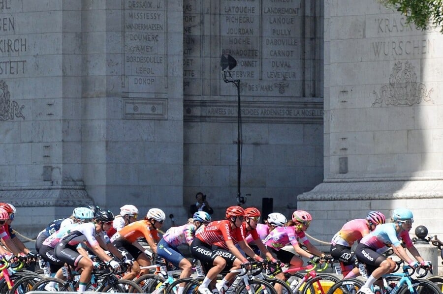 Kolarze biorący udział w wyścigu Tour de France Femmes jadący w zwartej grupie ulicami Paryża na tle Łuku Triumfalnego.