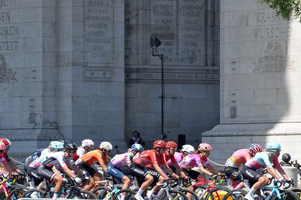 Kolarze biorący udział w wyścigu Tour de France Femmes jadący w zwartej grupie ulicami Paryża na tle Łuku Triumfalnego.