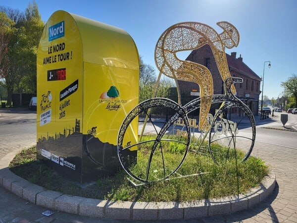 Na zdjęciu widoczna jest efektowna rzeźba kolarza oraz żółta instalacja promująca Tour de France 2025 w miejscowości Lauwin-Planque. Elementy dekoracyjne nawiązują do prestiżowego wyścigu i podkreślają zaangażowanie regionu Nord w organizację wydarzenia. Całość ustawiona jest na rondzie, a w tle widać zabudowę i zieleń, co tworzy wyjątkowy klimat sportowego święta.