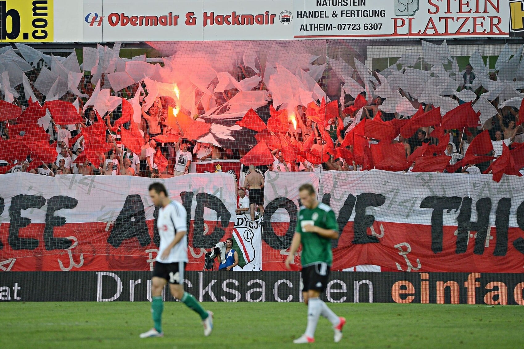 Oprawa kibiców Legii Warszawa z sektorówką i racami podczas meczu piłkarskiego – czerwono-białe flagi oraz transparent na trybunach, hołd patriotyczny na stadionie.