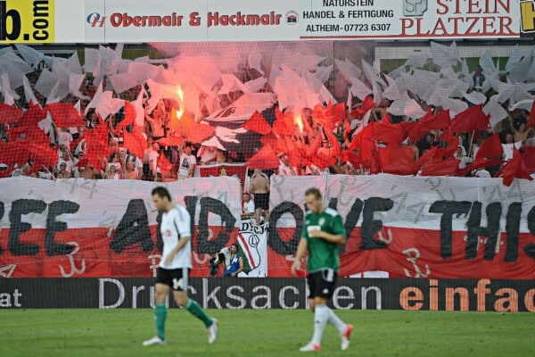 Oprawa kibiców Legii Warszawa z sektorówką i racami podczas meczu piłkarskiego – czerwono-białe flagi oraz transparent na trybunach, hołd patriotyczny na stadionie.