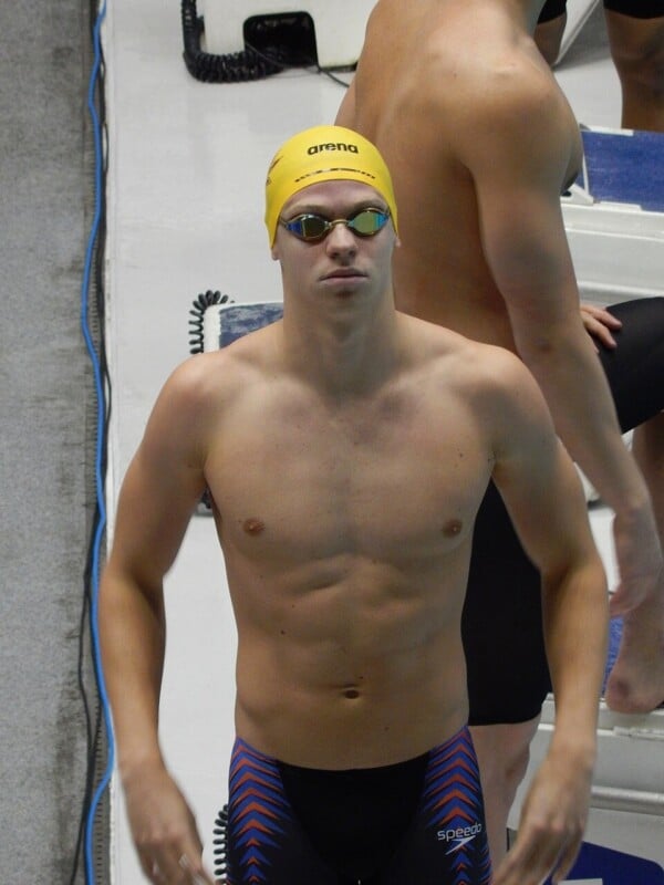 Leon Marchand w żółtym czepku Arena i stroju Speedo na starcie pływackim, przygotowujący się do wyścigu na basenie.