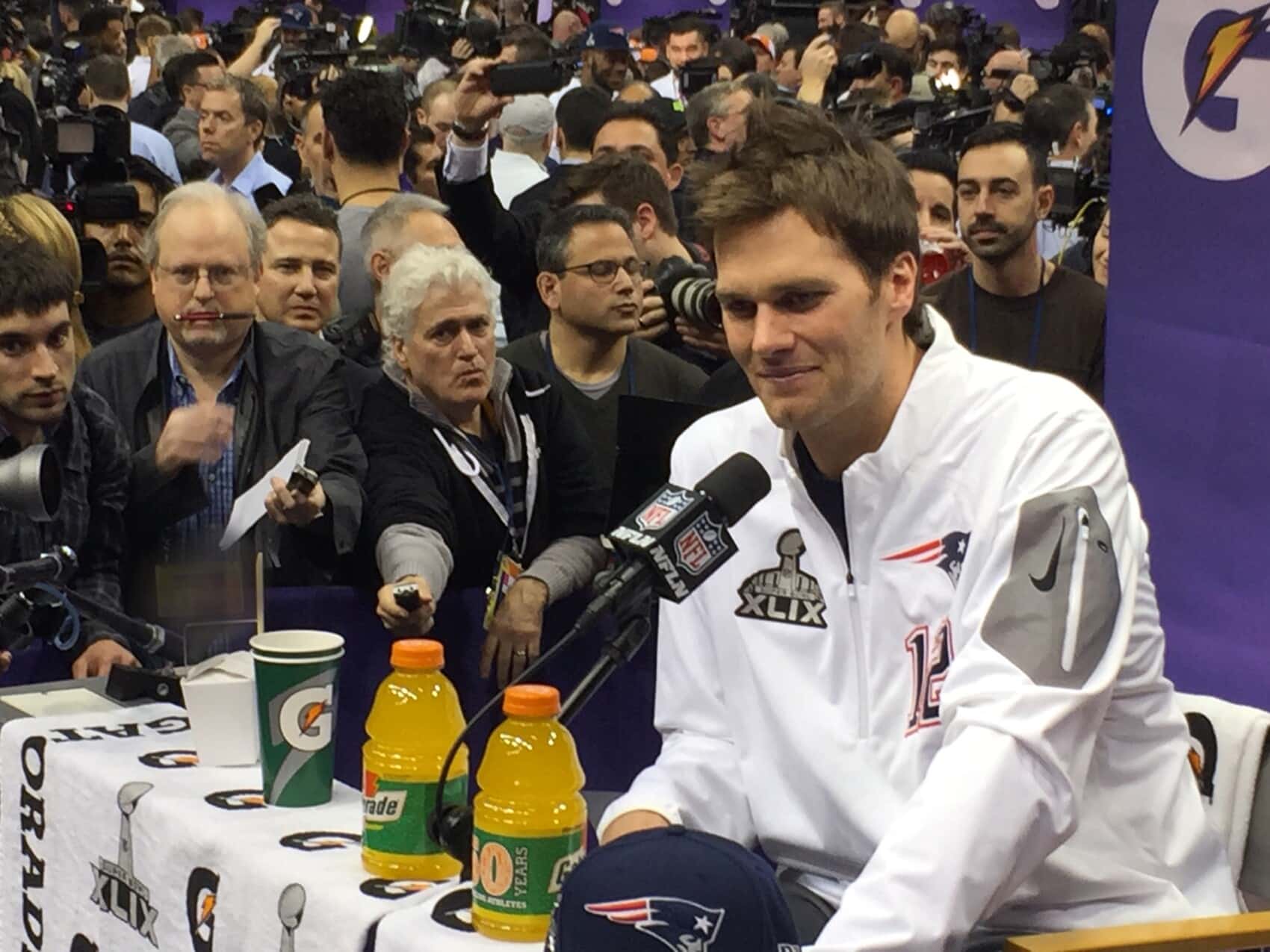 Konferencja prasowa NFL Super Bowl XLIX – zawodnik New England Patriots w białej kurtce, mikrofon NFL Network, napoje Gatorade i tłum fotoreporterów.
