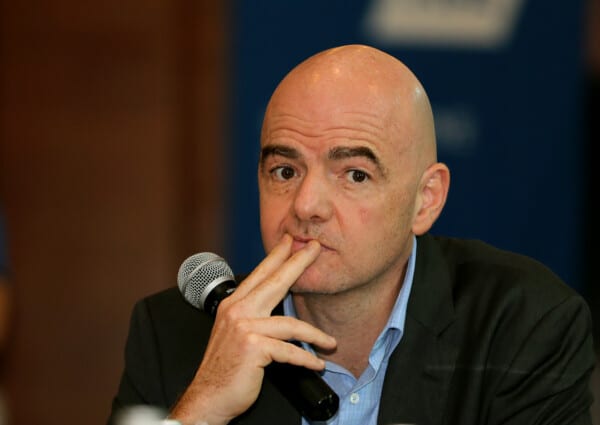 Gianni Infantino przemawia do mikrofonu na oficjalnej konferencji, ubrany w garnitur, tło z logotypem FIFA.