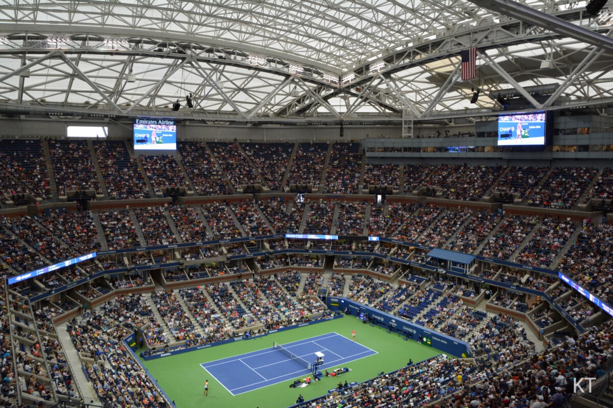 Wnętrze stadionu Arthur Ashe z zamkniętym dachem, widoczne pełne trybuny kibiców oraz kort podczas meczu tenisowego US Open