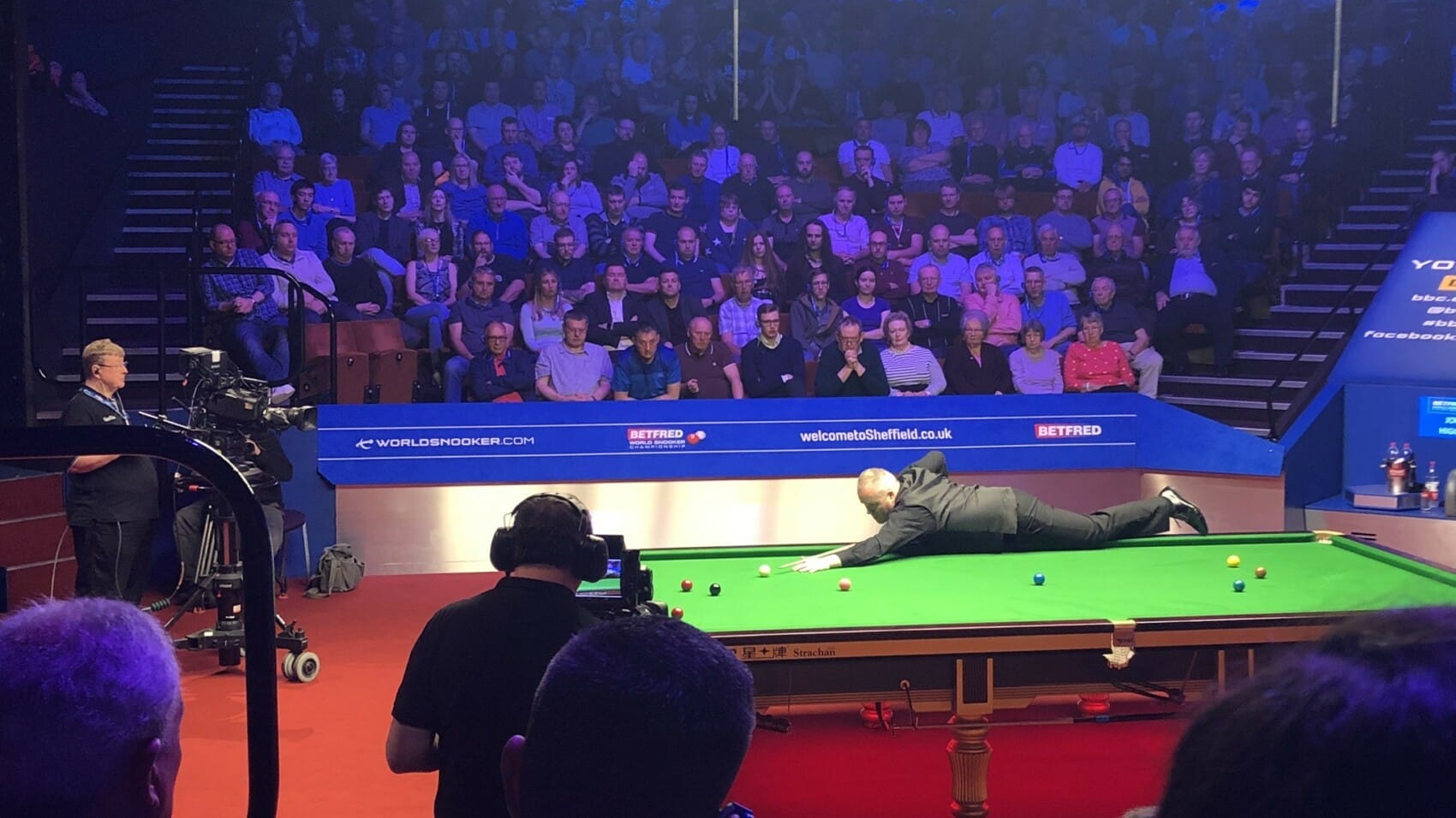 John Higgins wykonuje uderzenie przy stole podczas profesjonalnego meczu snookera w Crucible Theatre w Sheffield, 2019 rok, z widownią i ekipą telewizyjną w tle.