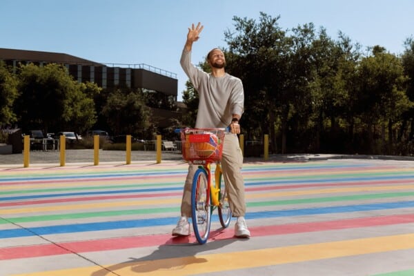 Stephen Curry jedzie kolorowym rowerem Google po tęczowej, wielobarwnej ulicy, w casualowym stroju, podczas słonecznego dnia.