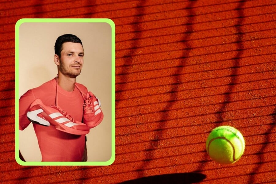 Polski tenisista w czerwonym stroju z parą czerwonych butów sportowych Adidas na szyi, na tle kortu ziemnego z żółtą piłką tenisową.
