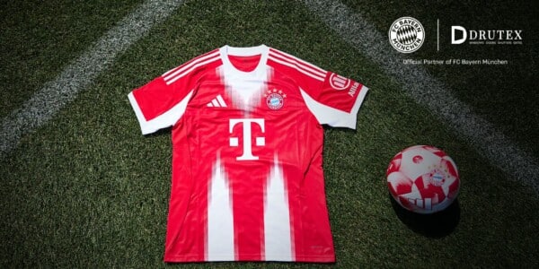 Koszulka domowa FC Bayern Monachium leżąca na murawie boiska wraz z piłką klubową, widoczne logo sponsora DRUTEX i T-Mobile oraz herb klubu.