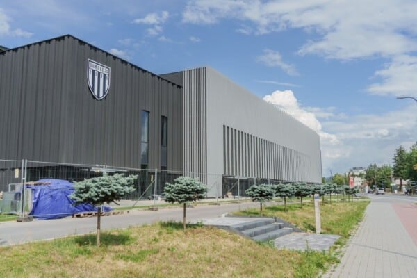 Nowoczesny stadion Sandecji Nowy Sącz z charakterystyczną czarną fasadą i logo klubu, widok od ulicy, pośród młodych drzewek i w słoneczny dzień.