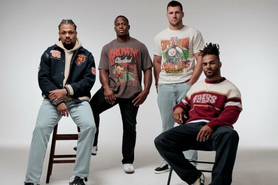 Grupa czterech mężczyzn w modnych ubraniach typu vintage z motywami drużyn NFL, pozujących razem w stylowym studio na neutralnym tle.