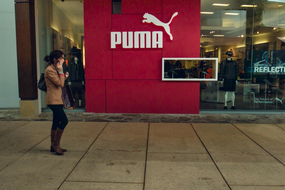 Kobieta idąca przed wejściem do sklepu Puma z charakterystycznym czerwonym panelem i dużym logo marki na tle witryny sklepowej