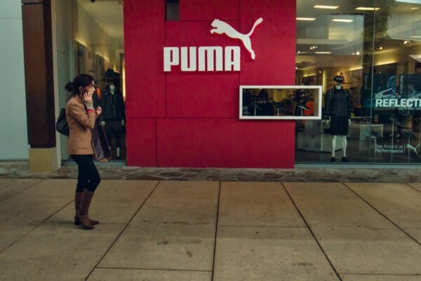 Kobieta idąca przed wejściem do sklepu Puma z charakterystycznym czerwonym panelem i dużym logo marki na tle witryny sklepowej