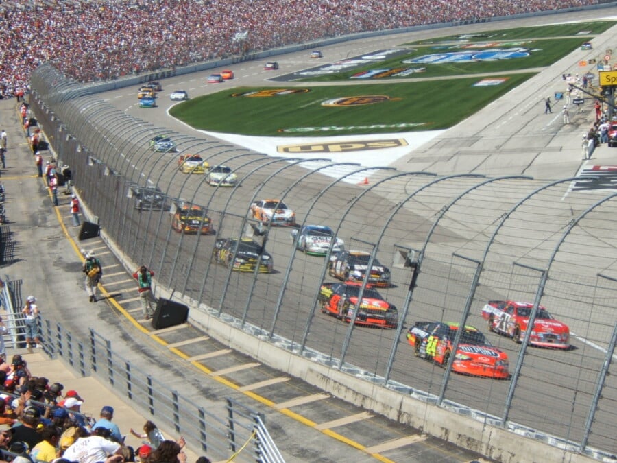 Wyścig NASCAR na zatłoczonym stadionie z kolorowymi samochodami na torze i liczną publicznością na trybunach za ochronną siatką.