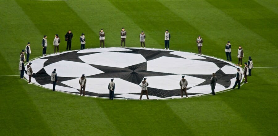 Champions League star logo displayed on green football pitch with officials in formal wear surrounding the iconic UEFA symbol during pre-match ceremony