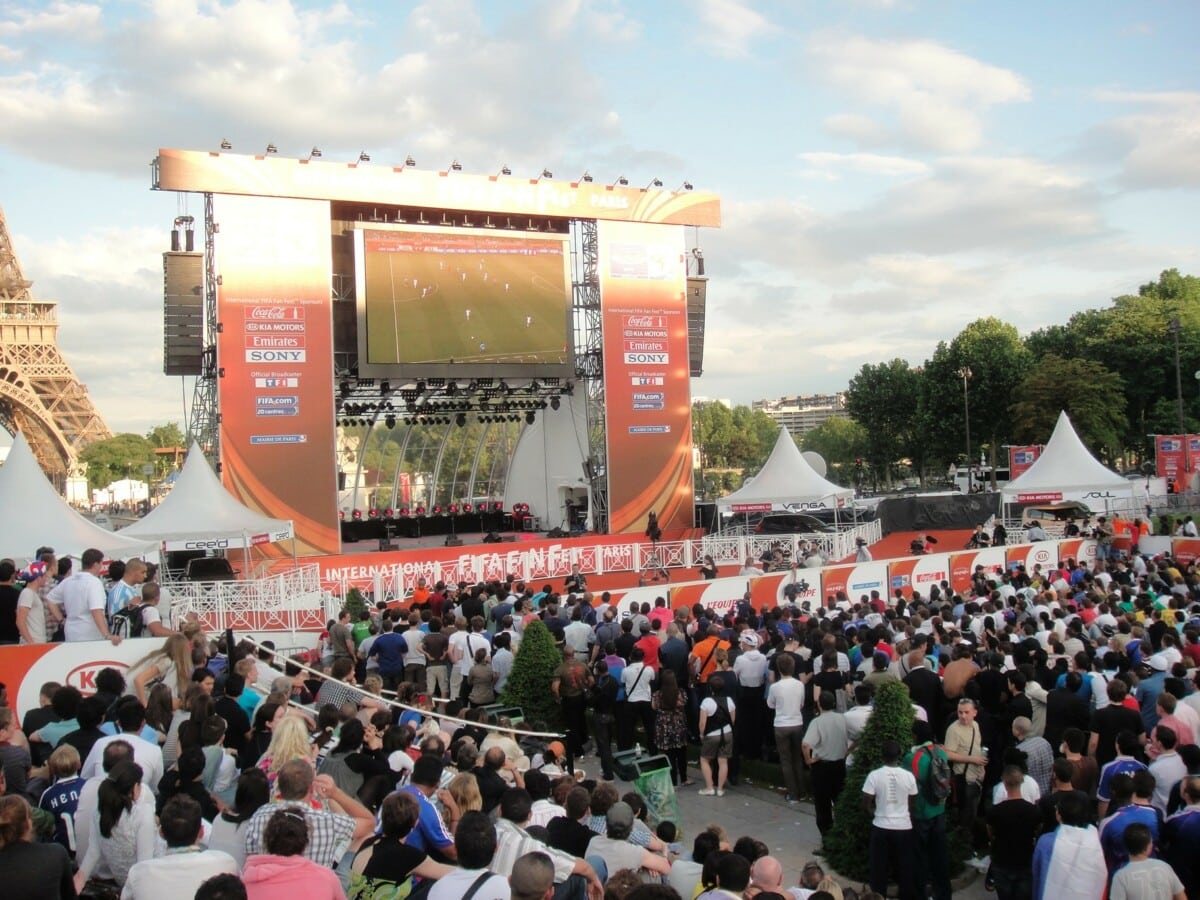 Tłum kibiców ogląda mecz piłkarski na dużym ekranie podczas FIFA Fan Fest w Paryżu, na tle Wieży Eiffla, w słoneczny dzień