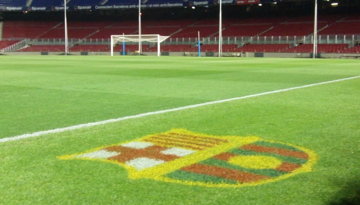 Boisko na stadionie Camp Nou z widocznym herbem FC Barcelona na trawie, pustymi trybunami i bramką w tle