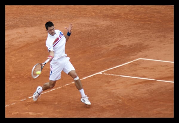 Novak Djoković grający mecz tenisa na korcie ziemnym, wykonuje forehand w białym stroju sportowym