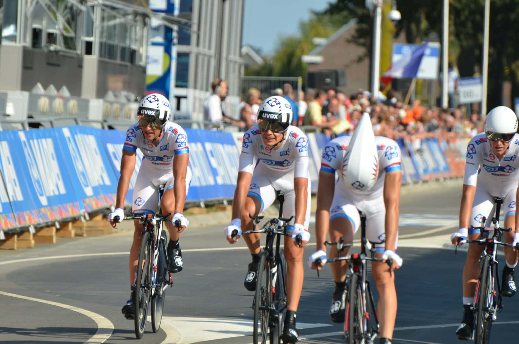 Czterech kolarzy zespołu FDJ w białych strojach i kaskach aerodynamicznych bierze udział w drużynowej jeździe na czas podczas profesjonalnego wyścigu kolarskiego, przy barierkach z kibicami w tle.