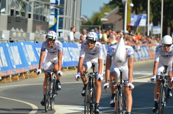 Czterech kolarzy zespołu FDJ w białych strojach i kaskach aerodynamicznych bierze udział w drużynowej jeździe na czas podczas profesjonalnego wyścigu kolarskiego, przy barierkach z kibicami w tle.