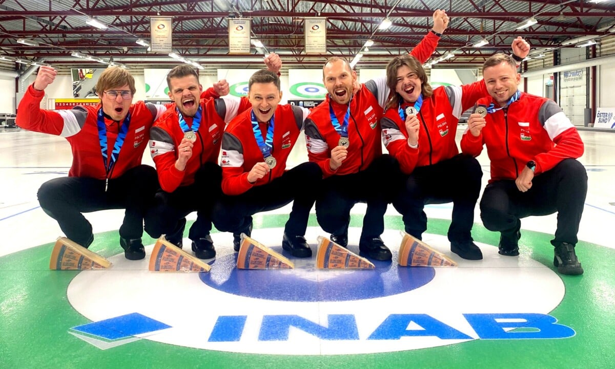 Reprezentacja Polski w curlingu pozująca na lodowisku z medalami po wygranym turnieju, ubrani w czerwone stroje z napisem Polska.