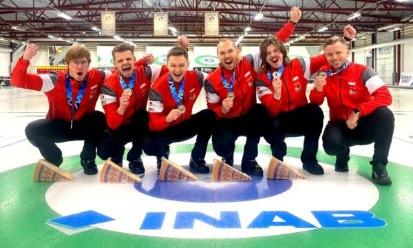 Reprezentacja Polski w curlingu pozująca na lodowisku z medalami po wygranym turnieju, ubrani w czerwone stroje z napisem Polska.