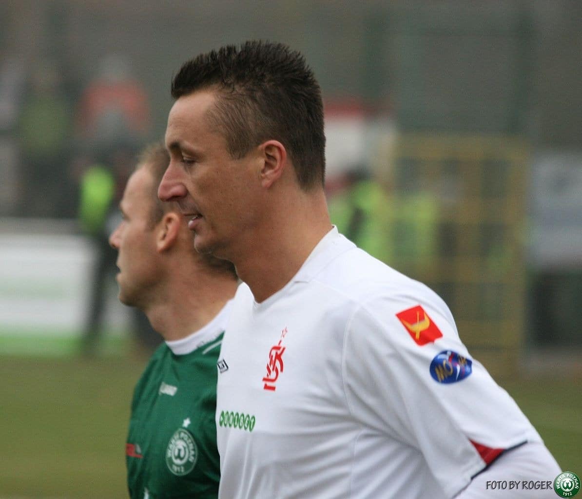 Zawodnik ŁKS Łódź w białej koszulce z czerwonym herbem klubu na piersi oraz naszywkami sponsorskim i miejskim na rękawie, stojący obok piłkarza Warty Poznań w zielonym stroju podczas meczu piłki nożnej.