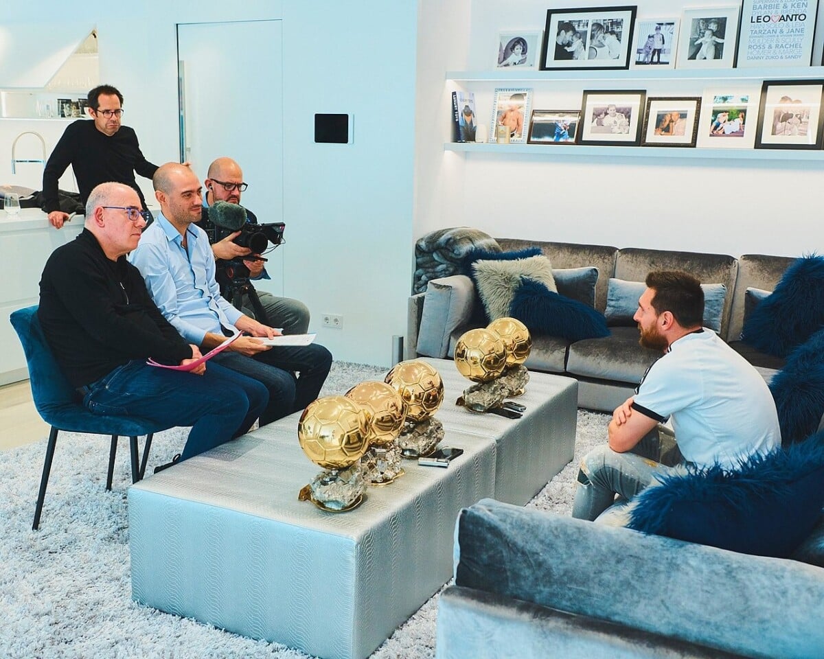 Sześć Złotych Piłek na stoliku w eleganckim salonie. Kilka osób siedzi i rozmawia przy ekskluzywnej kanapie w otoczeniu pamiątkowych fotografii.