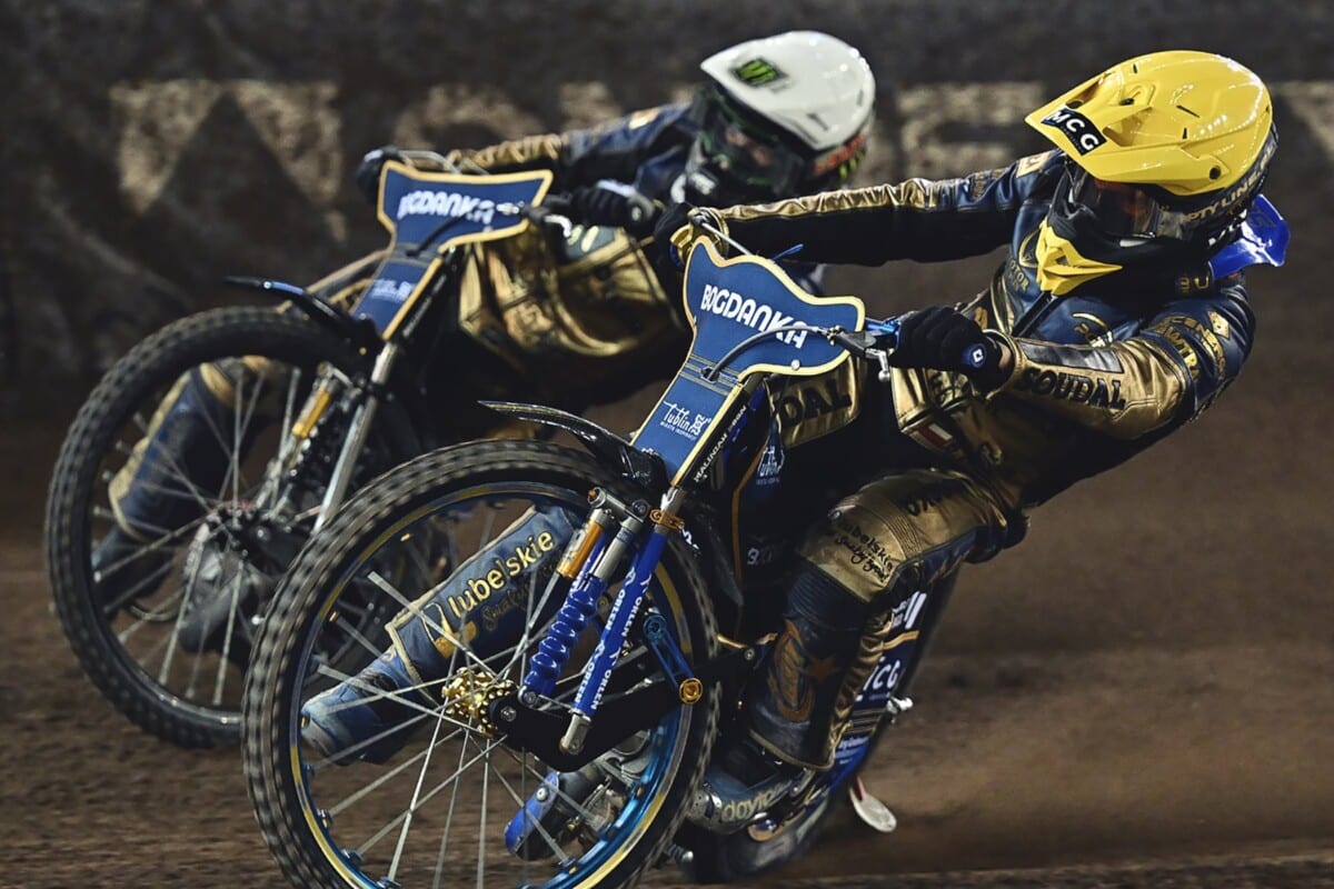 Dwóch zawodników podczas dynamicznego wyścigu żużlowego na torze w Toruniu, w żółtym i białym kasku.