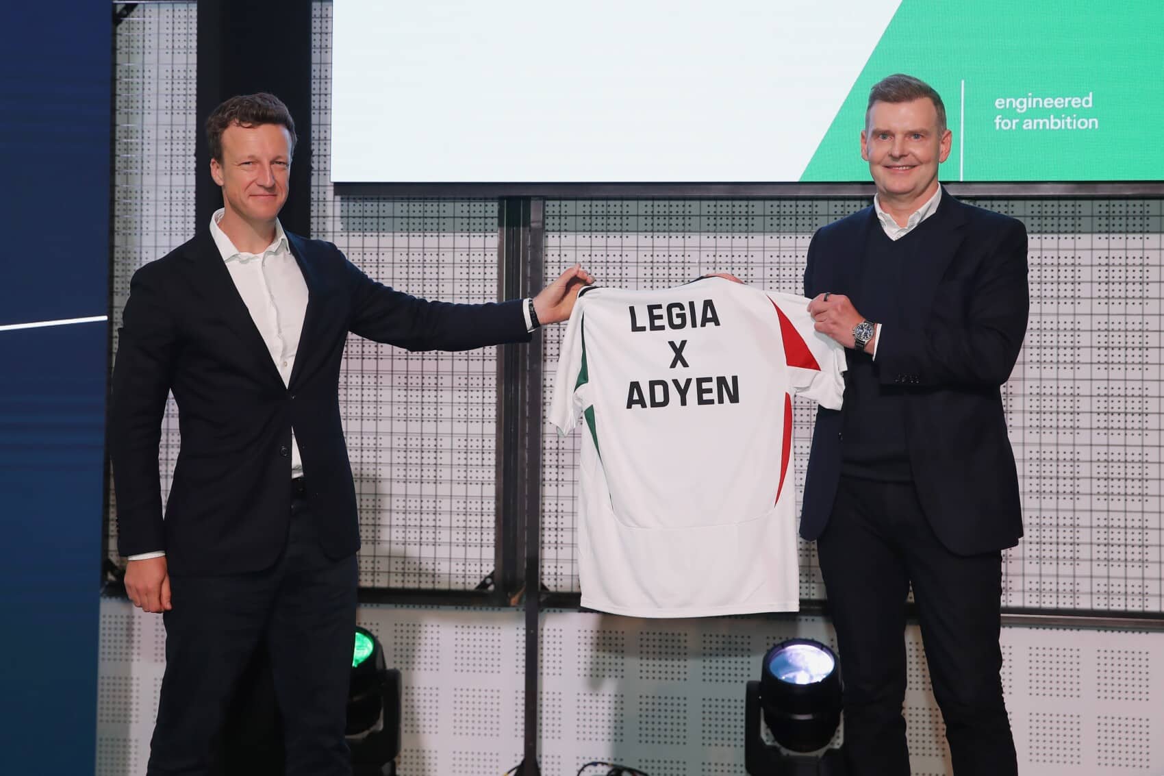 Dwóch mężczyzn w garniturach prezentuje koszulkę piłkarską z napisem LEGIA X ADYEN podczas oficjalnego wydarzenia partnerskiego.