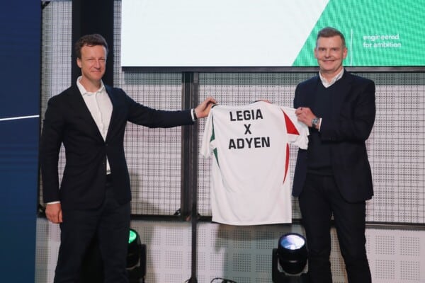 Dwóch mężczyzn w garniturach prezentuje koszulkę piłkarską z napisem LEGIA X ADYEN podczas oficjalnego wydarzenia partnerskiego.