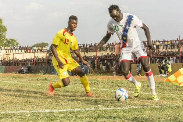 Piłkarz reprezentacji Sudanu Południowego z piłką przy nodze walczy o pozycję z rywalem w żółtym stroju podczas meczu na trawiastym boisku, w tle tłum kibiców na trybunach.