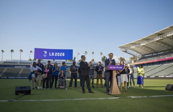 Grupa organizatorów i sportowców na murawie stadionu podczas prezentacji igrzysk LA 2028, z mównicą „Follow the Sun” i dużym ekranem z logo LA 2028 w tle.