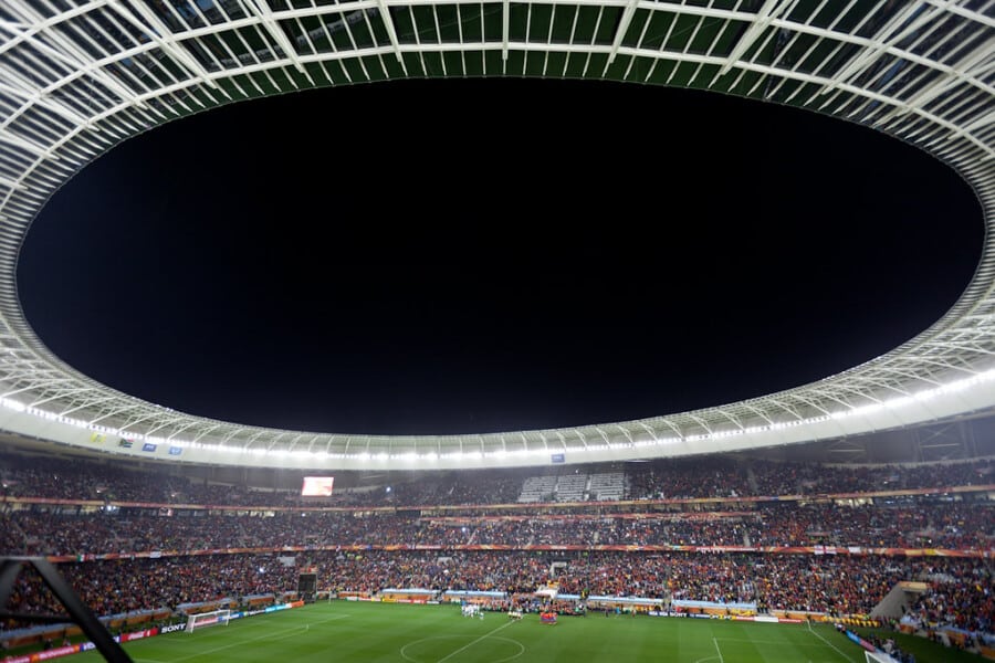 Panoramiczny widok nocnego meczu piłkarskiego na Cape Town Stadium w Kapsztadzie, z pełnymi trybunami kibiców i jasno oświetlonym owalnym dachem nad murawą.