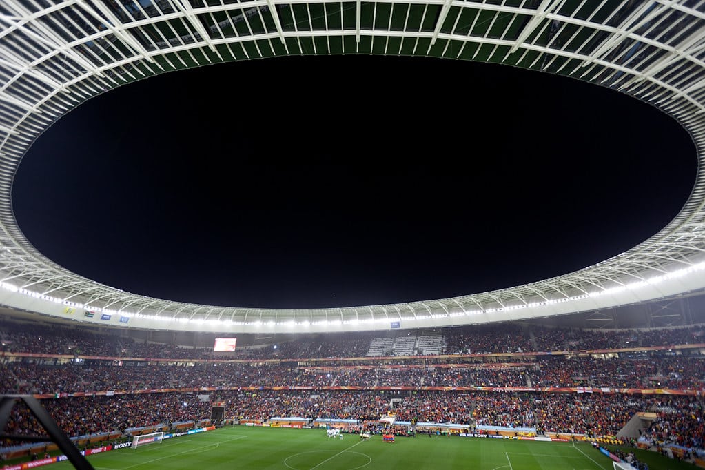 Panoramiczny widok nocnego meczu piłkarskiego na Cape Town Stadium w Kapsztadzie, z pełnymi trybunami kibiców i jasno oświetlonym owalnym dachem nad murawą.
